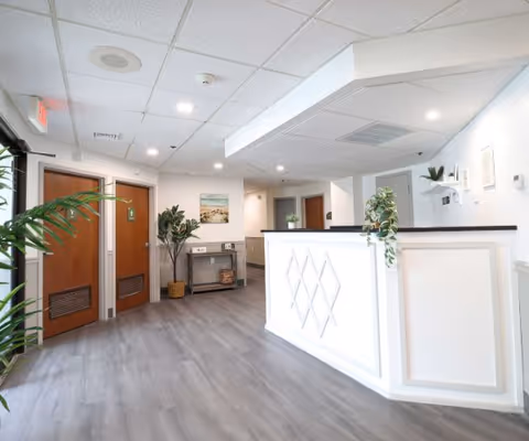 Bright and clean reception area with a white front desk featuring diamond patterns, light gray wood flooring, white walls, and ceiling tiles with recessed lighting. There are two wooden doors on the left side, a small table with plants and decor against the wall, and green plants placed around the space.