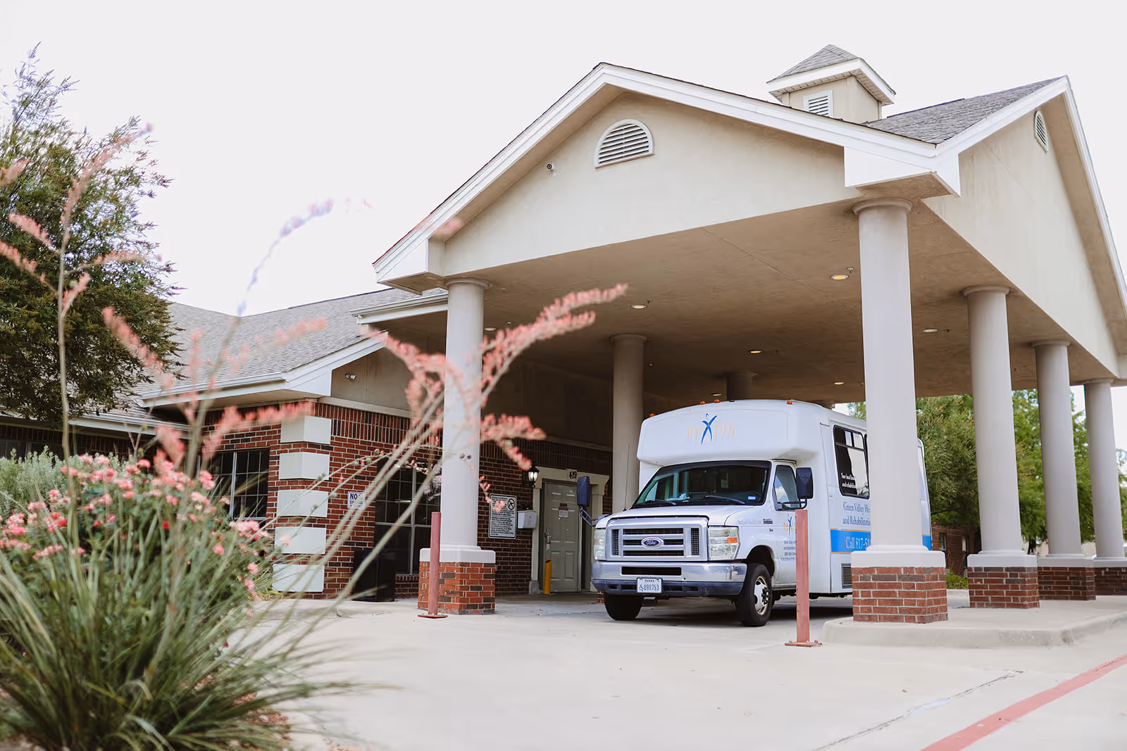 Covered entrance of a healthcare facility with a shuttle van parked under a large porte-cochère and plants in the foreground.