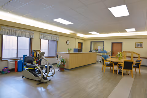 A spacious room with exercise equipment including a stationary bike and weights near two large windows. There is a reception desk or counter in the middle of the room, a round table with chairs, and some plants and decorations. The room has a light-colored floor and ceiling with fluorescent lighting.