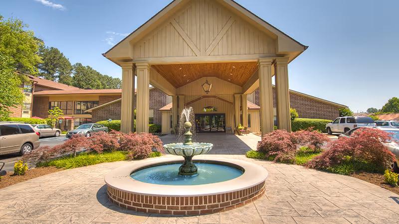 Front exterior view of Hickory Valley A Senior Living Community featuring a covered entrance with large columns, a decorative fountain in the foreground, landscaped bushes, and parked cars on either side.