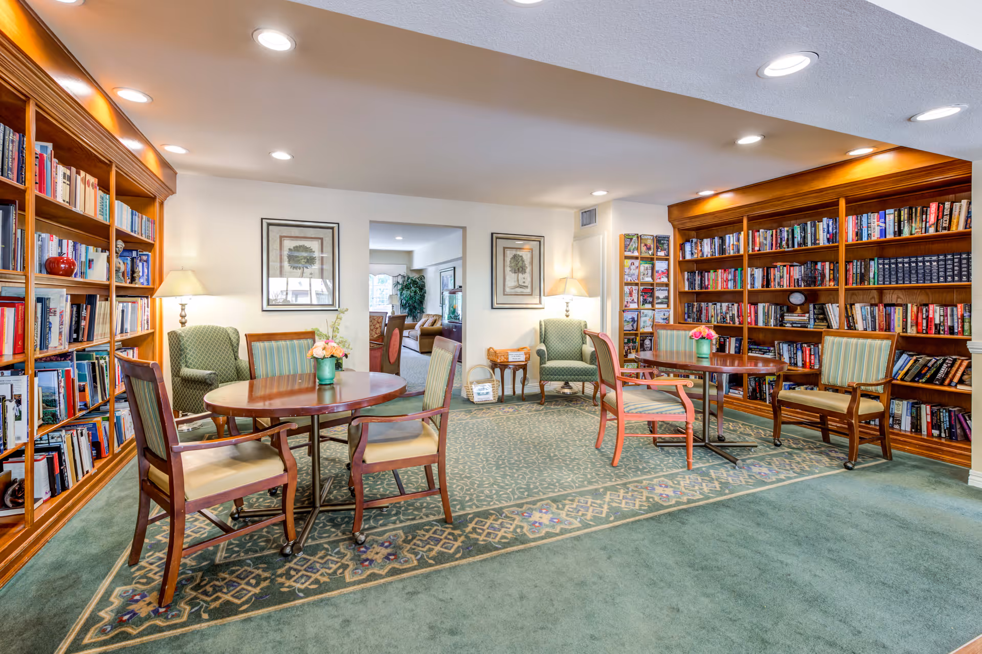 A cozy library room in a senior living facility with wooden bookshelves filled with books along two walls. There are two round wooden tables with chairs around them, a patterned green carpet, two green upholstered armchairs with lamps behind them, and framed artwork on the walls. The room is well-lit with recessed ceiling lights.