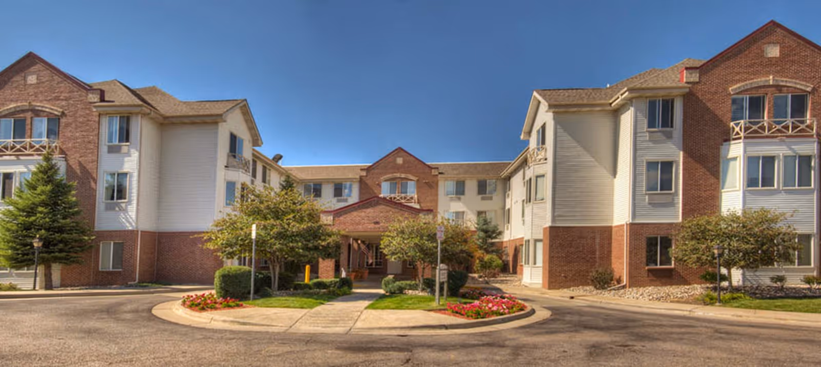 Exterior view of a three-story assisted living facility building with a combination of brick and white siding. The entrance is centered with a covered porch, surrounded by landscaped greenery and flower beds. The sky is clear and blue.