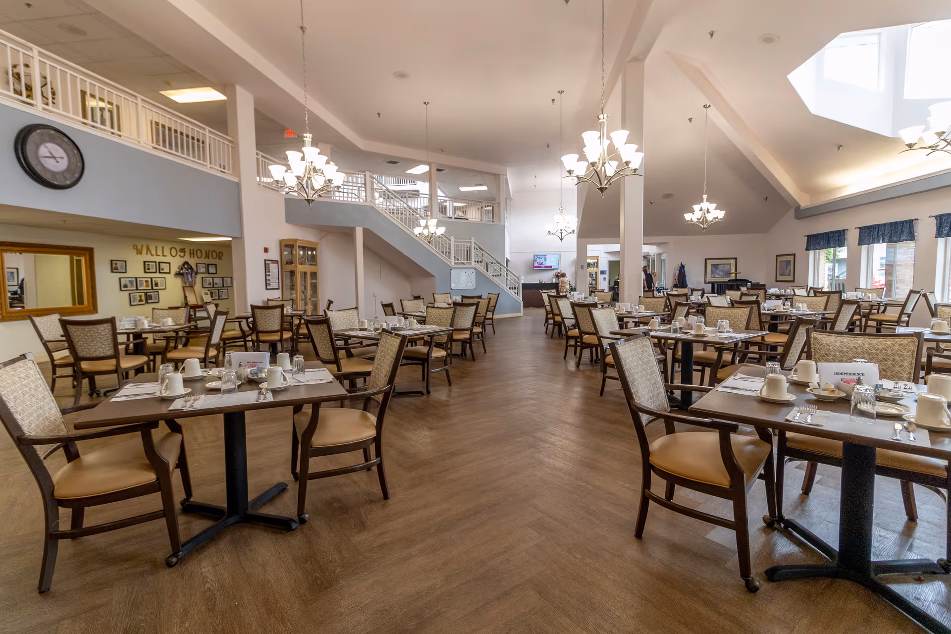 Spacious dining room with multiple tables and chairs arranged neatly. Each table is set with cups, glasses, and utensils. The room features chandeliers hanging from the ceiling, a staircase leading to an upper level, and large windows allowing natural light to enter. A clock and a 'Wall of Honor' display are visible on one wall.