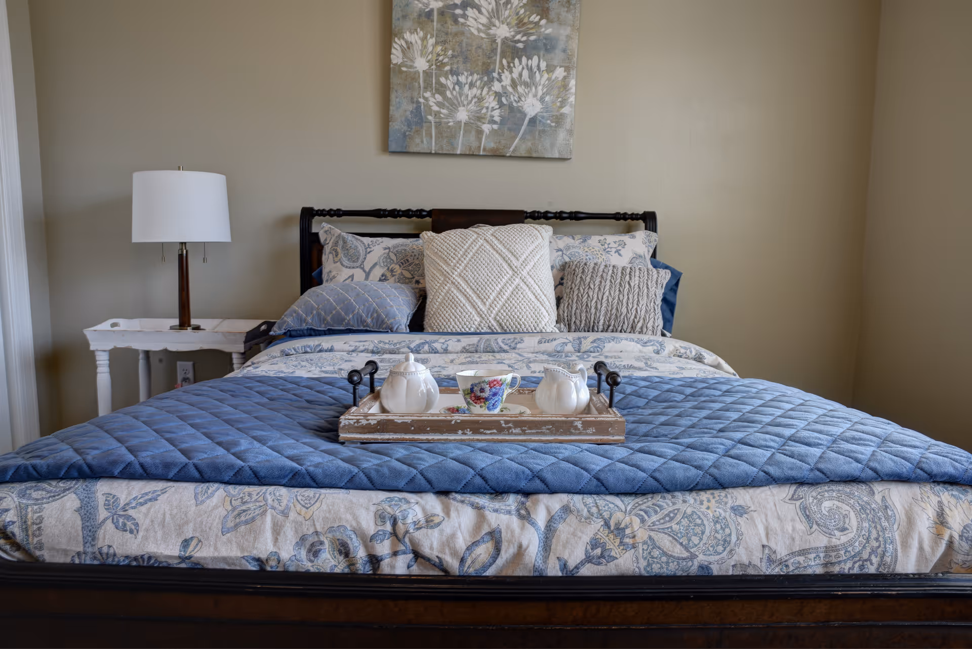 A neatly made bed with a blue quilted blanket and patterned sheets, adorned with several decorative pillows. A wooden tray with a teapot, creamer, and a floral teacup sits on the bed. To the left of the bed is a white side table with a lamp. A floral painting hangs on the beige wall above the bed.