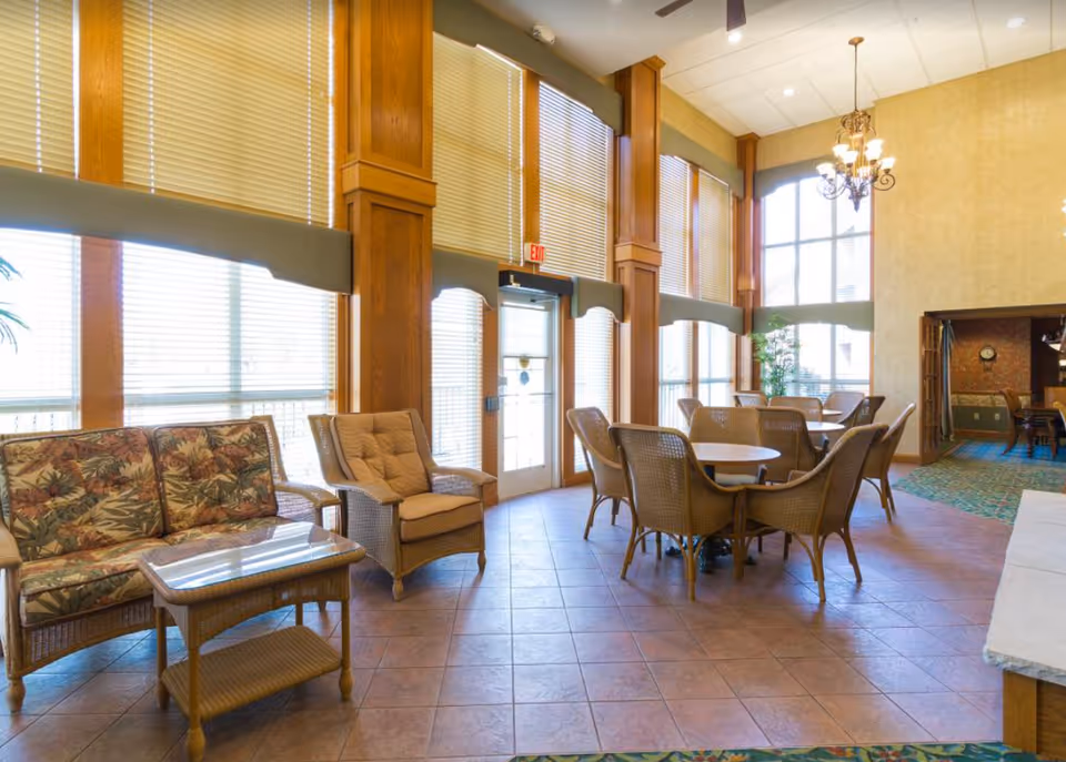 A bright and spacious common area with large windows covered by blinds, featuring wicker furniture including cushioned chairs, a loveseat with floral upholstery, and round tables with chairs. The room has tiled flooring, wooden columns, and a chandelier hanging from the ceiling.