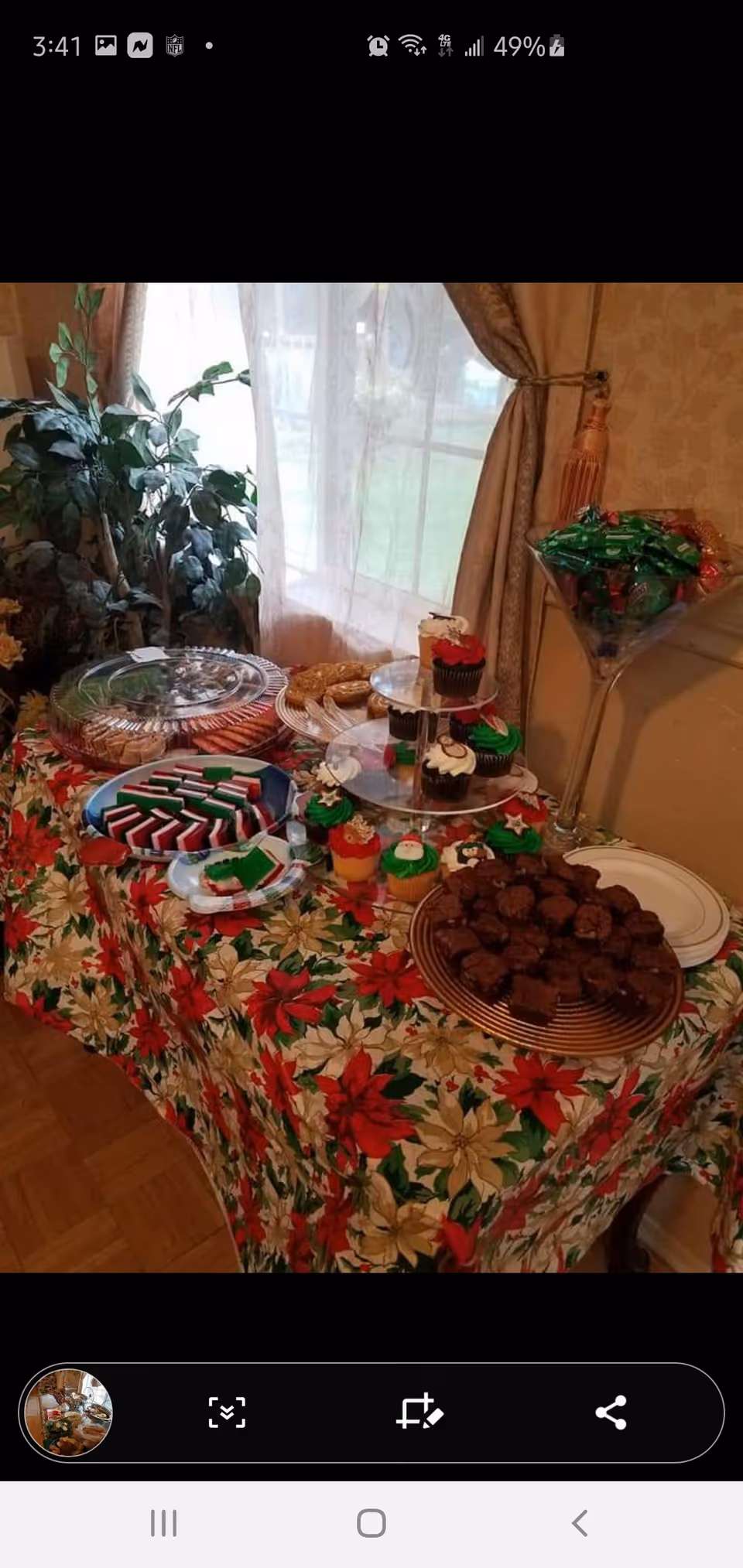 A festive table covered with a poinsettia-patterned tablecloth displaying an assortment of holiday-themed treats including cupcakes decorated with Christmas designs, brownies, and other cookies. The table is positioned near a window with sheer curtains and a potted plant is visible in the background.