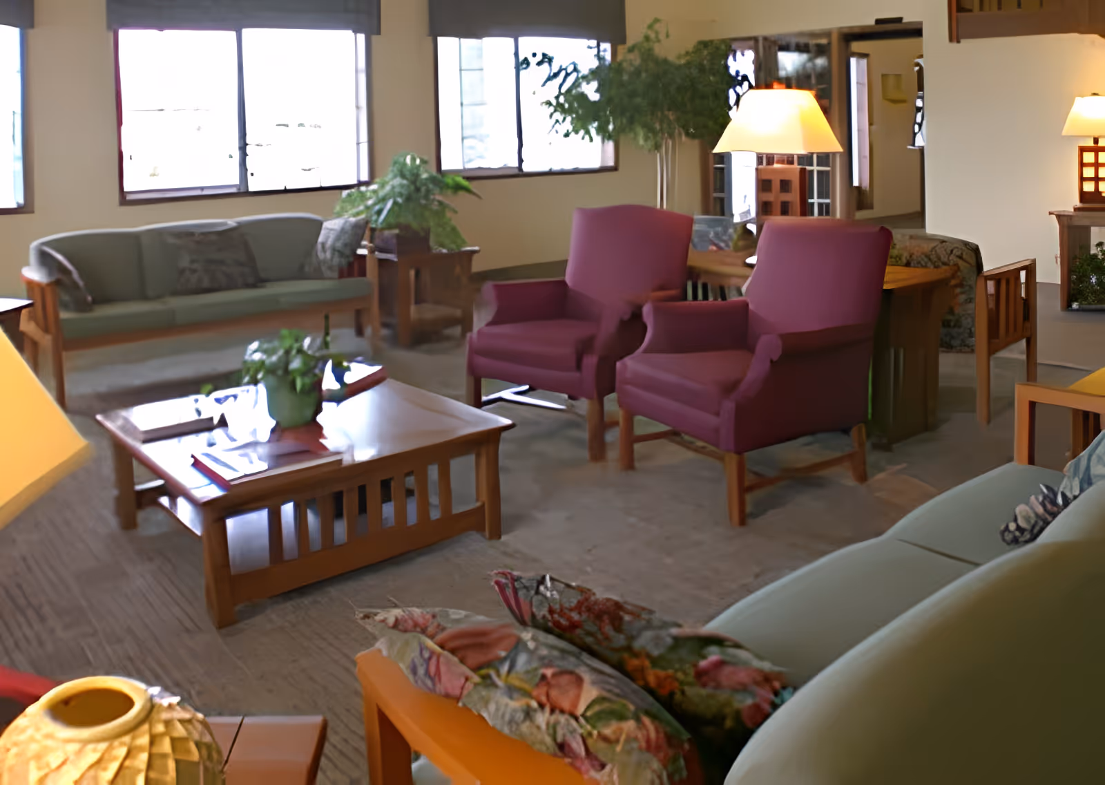 A cozy living room area with multiple seating options including green sofas and two purple armchairs. There are wooden coffee tables with plants and books on them, several table lamps providing warm lighting, and large windows letting in natural light. The room has a calm and inviting atmosphere with neutral-colored walls and carpeted floor.