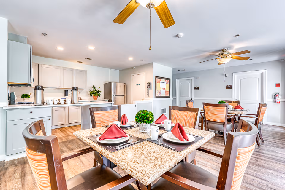 A bright dining area in a senior living facility with multiple tables set with plates, cups, and red folded napkins. The room features wooden chairs with striped cushions, ceiling fans with lights, and a kitchen area in the background with white cabinets, a stainless steel refrigerator, and coffee dispensers on the counter.