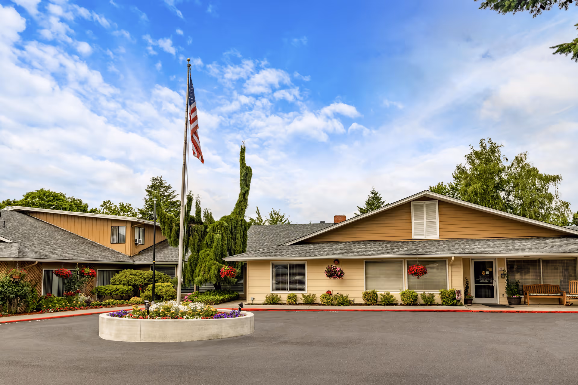 Exterior view of Kamlu Retirement Inn-Vancouver showing a single-story building with beige siding and a gray roof. There is a circular flower bed with colorful flowers and an American flagpole in the center of the paved driveway. The building is surrounded by green trees and hanging flower baskets, with a bench near the entrance.