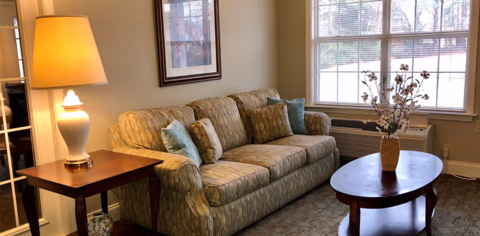 A cozy living room area with a patterned beige sofa adorned with multiple cushions, a wooden side table with a white lamp, a wooden coffee table with a vase of flowers, and a large window letting in natural light.