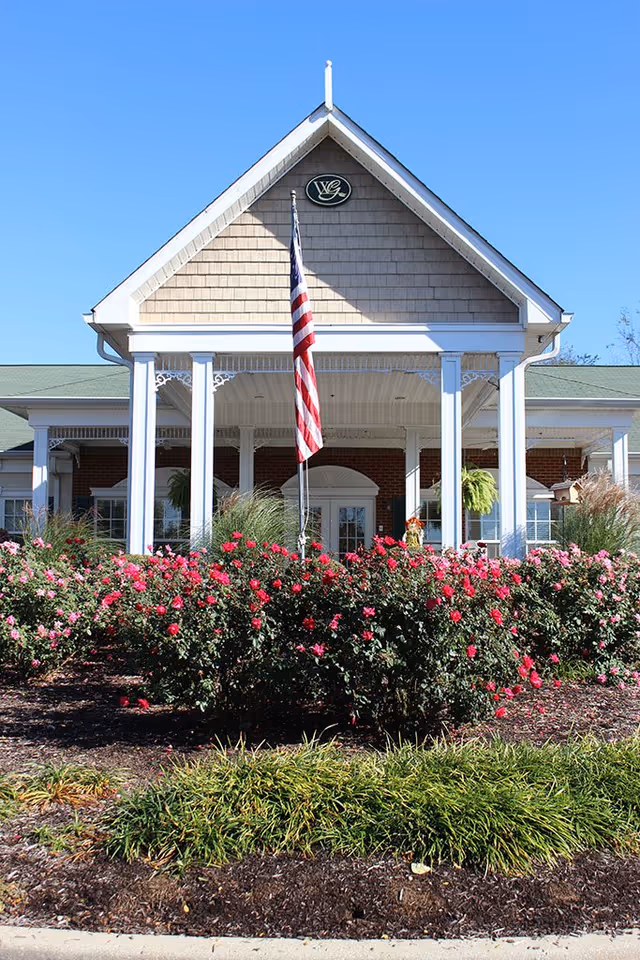 Front exterior view of a senior living facility building with a peaked roof, white columns, an American flag in front, and a garden with blooming pink and red flowers.