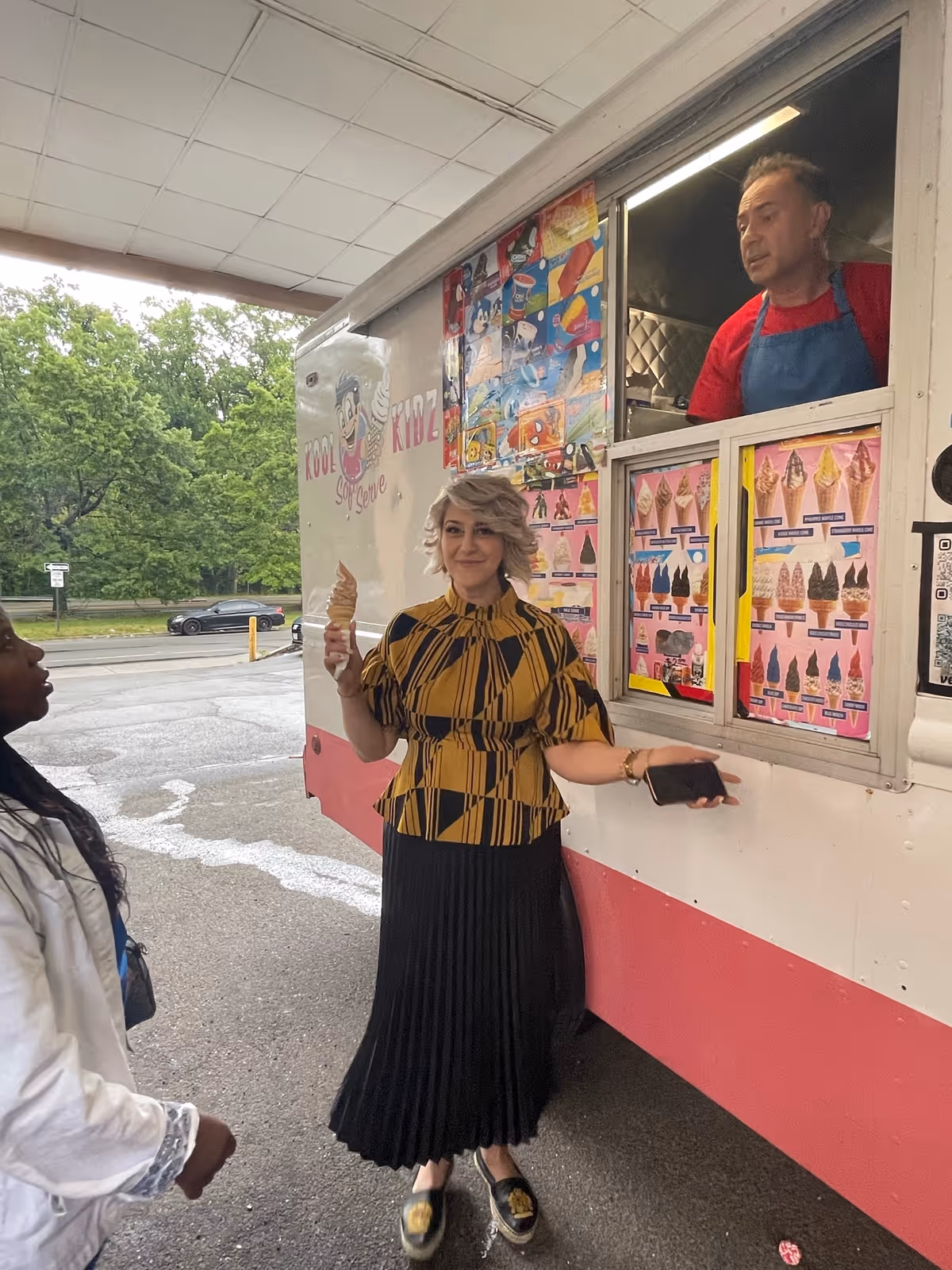 A woman holding an ice cream cone stands in front of an ice cream truck, smiling. Another person is partially visible on the left side of the image. A man wearing a red shirt and blue apron is inside the truck serving ice cream. The background shows trees and a parked car.