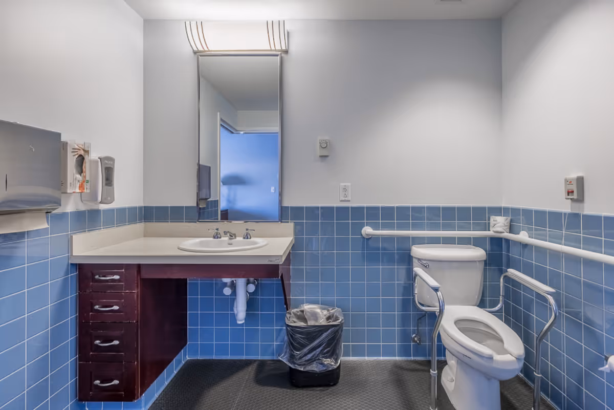 A clean and accessible bathroom with blue tiled walls and white upper walls. The bathroom features a white sink with a countertop and dark wood drawers underneath, a large mirror above the sink, a toilet with support rails on both sides, a trash can, a paper towel dispenser, and a soap dispenser mounted on the wall.