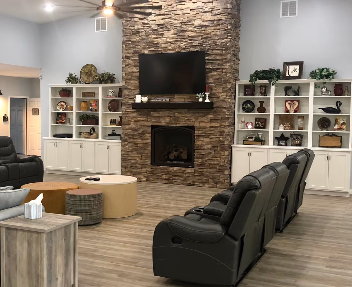 A cozy living room area with a stone fireplace and a mounted flat-screen TV above it. The room features white built-in shelves on either side of the fireplace, decorated with various plants, vases, and decorative items. In front of the fireplace are three black leather recliners arranged in a row, along with a round coffee table and additional seating. The floor is wood, and the walls are painted light gray.