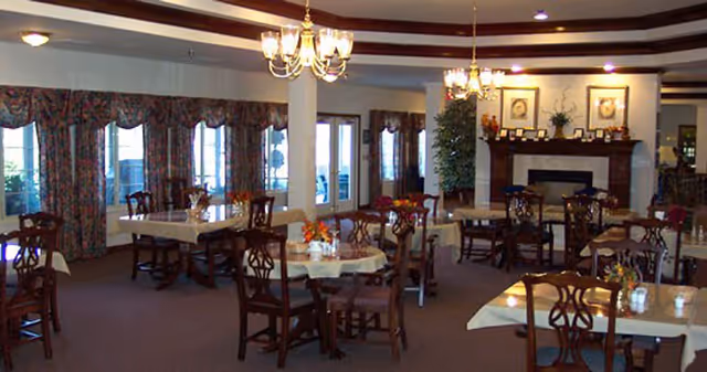 A spacious dining room with multiple wooden tables and chairs arranged neatly. The tables are covered with white tablecloths and decorated with small floral centerpieces. Large windows with patterned curtains allow natural light to fill the room. Two chandeliers hang from the ceiling, and there is a fireplace with framed artwork and decorative items on the mantel. The room has a warm and inviting atmosphere.