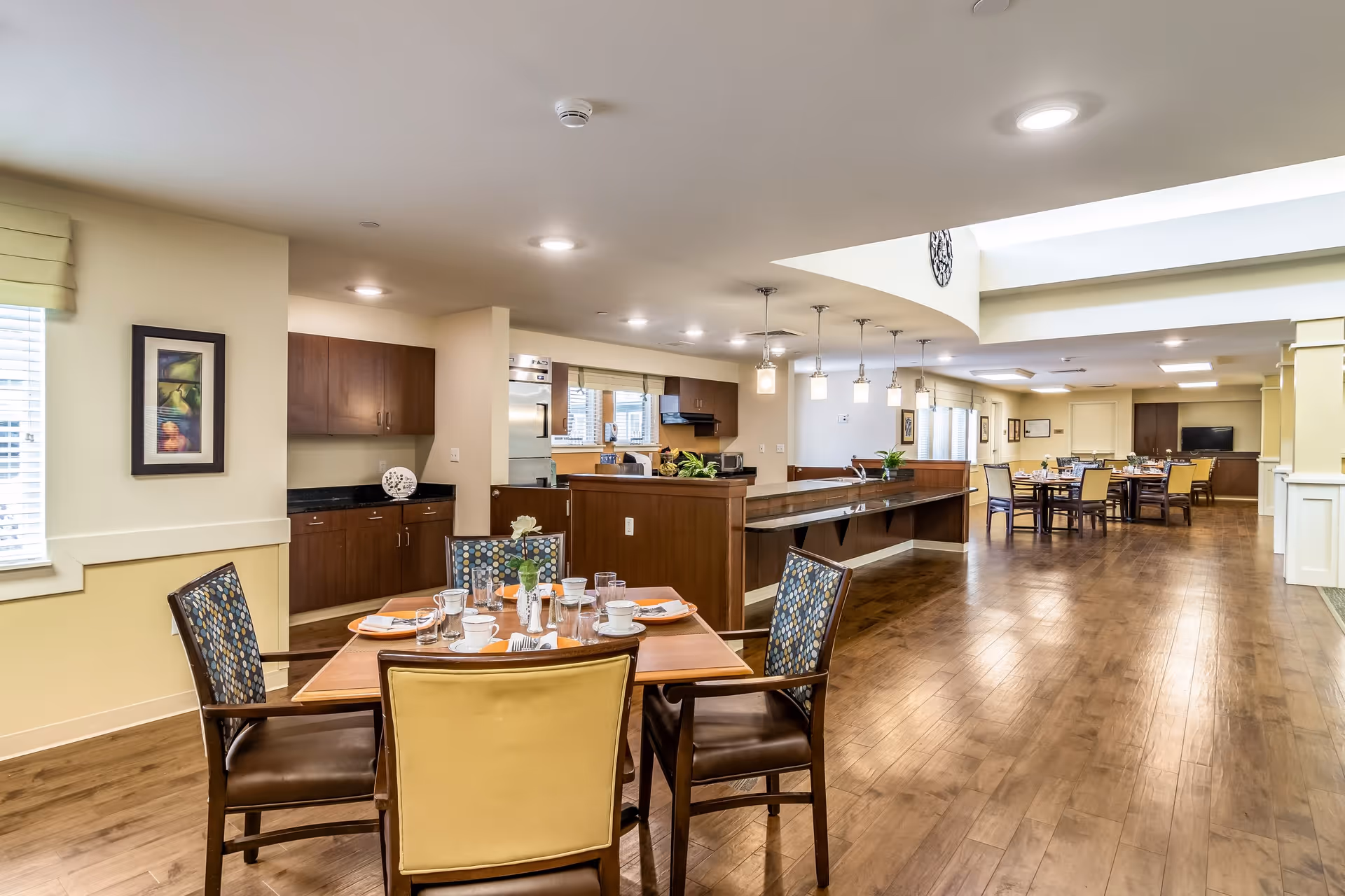 A spacious dining area in a senior living facility with wooden floors and multiple tables set for meals. The foreground shows a table with four chairs, place settings, and a small flower vase. The background includes a kitchen area with cabinets, countertops, and hanging pendant lights, as well as additional dining tables and chairs further back.