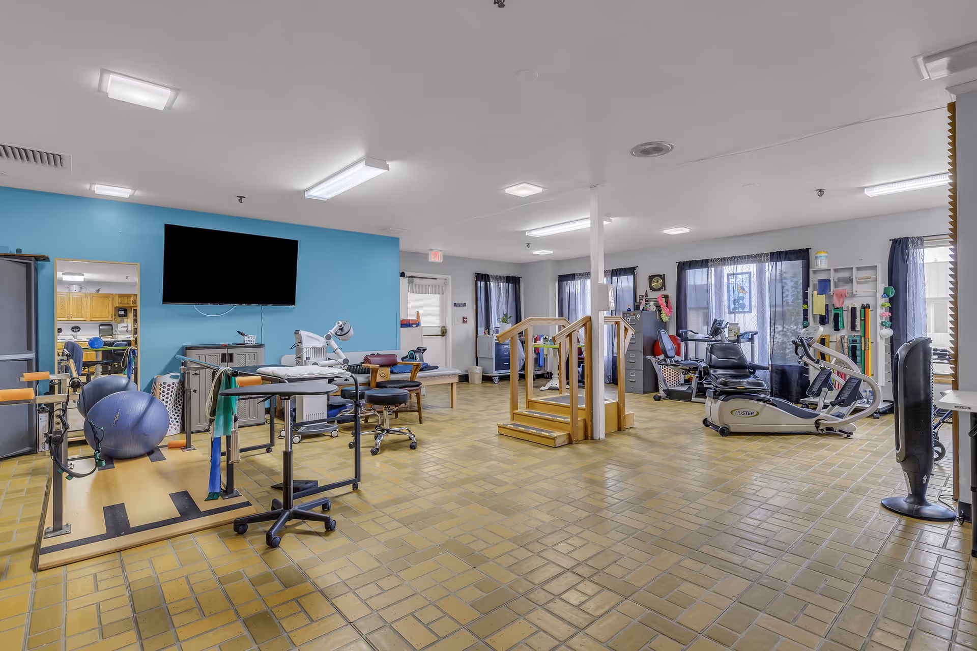 A spacious rehabilitation room with exercise equipment including stationary bikes, a step platform with handrails, therapy balls, and various physical therapy tools. The room has a tiled floor, blue accent wall with a mounted TV, and large windows with dark curtains allowing natural light.