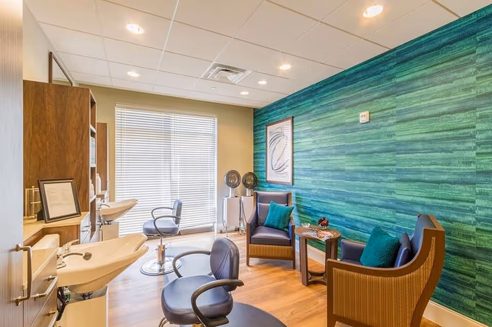 Interior view of a salon area in a senior living facility with two salon chairs in front of sinks, two armchairs with teal cushions, a small table with magazines, and a green textured accent wall.