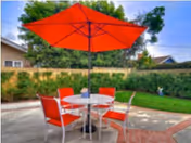 Outdoor patio area with a round table and four red chairs under a large red umbrella. The patio is surrounded by green grass, bushes, and trees, with neighboring houses visible in the background.