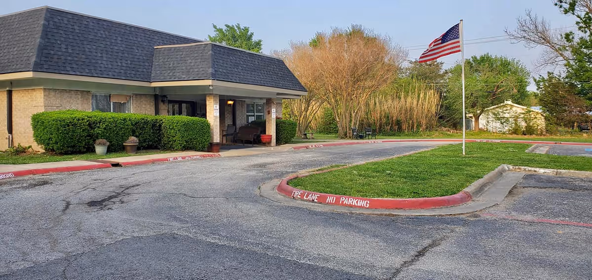 Exterior view of a single-story building with a dark shingled roof and beige brick walls, surrounded by green bushes. There is a paved driveway and parking area with red curbs marked 'FIRE LANE NO PARKING'. An American flag on a flagpole is positioned on a grassy island in the parking lot. Trees and a small white shed are visible in the background under a clear sky.