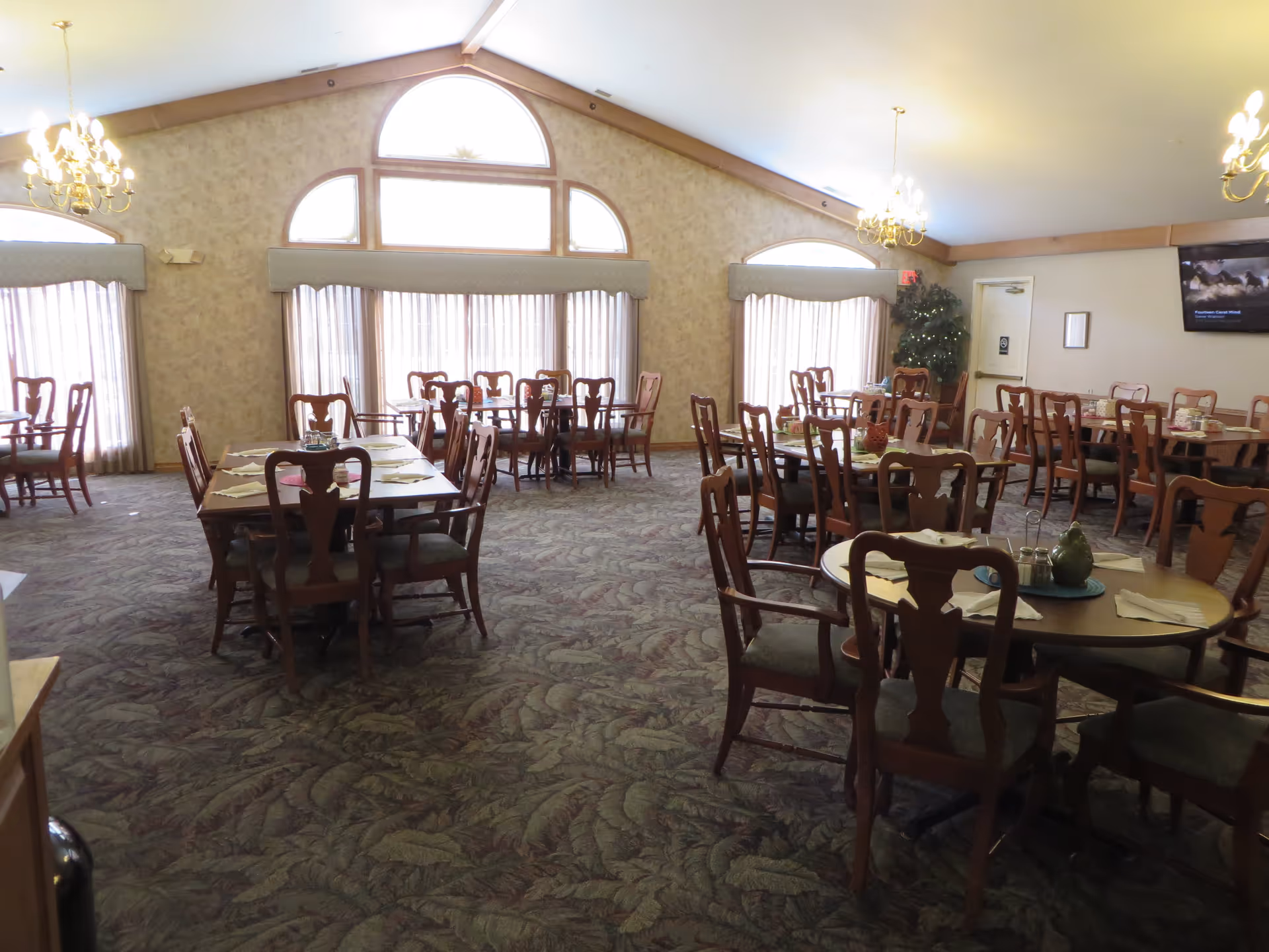 A spacious dining room with multiple wooden tables and chairs arranged neatly. The room features large windows with curtains, allowing natural light to enter. There are chandeliers hanging from the ceiling and a television mounted on the wall. The carpet has a leafy pattern, and the walls are decorated with light-colored wallpaper.