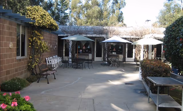 Outdoor courtyard patio with tables, umbrellas, chairs, benches, and plants in front of a single-story building.
