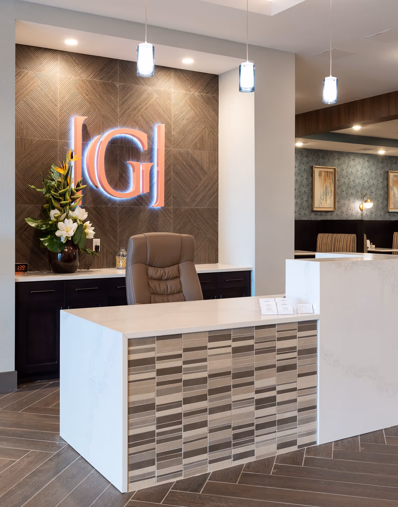 Reception desk area with a modern white and tiled front counter, a brown leather office chair, a decorative flower arrangement, and a back wall featuring a textured wood panel with a glowing 'G' logo. Pendant lights hang from the ceiling, and a seating area with framed artwork is visible in the background.