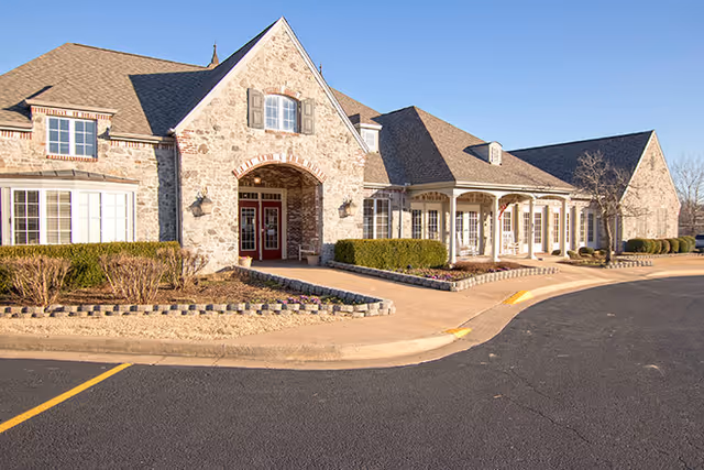 Exterior view of a stone building with a peaked roof and multiple windows, featuring a covered entrance and landscaped bushes along the front. The building is situated next to a paved driveway under a clear blue sky.