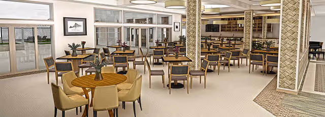 A spacious dining room with multiple round and square wooden tables surrounded by chairs. The room features large windows and glass doors on one side, allowing natural light to enter. There are decorative plants on some tables and framed artwork on the walls. The ceiling has modern circular light fixtures, and the floor is carpeted with a neutral tone.