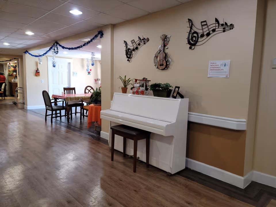 Interior view of a senior living facility hallway with a white upright piano against a beige wall decorated with musical note wall art and a guitar. There is a small brown bench in front of the piano. The hallway has wood flooring and ceiling lights. In the background, there is a dining area with a table covered with a striped tablecloth and several chairs. Blue and silver garlands hang from the ceiling, and small holiday decorations are visible on the walls and tables.