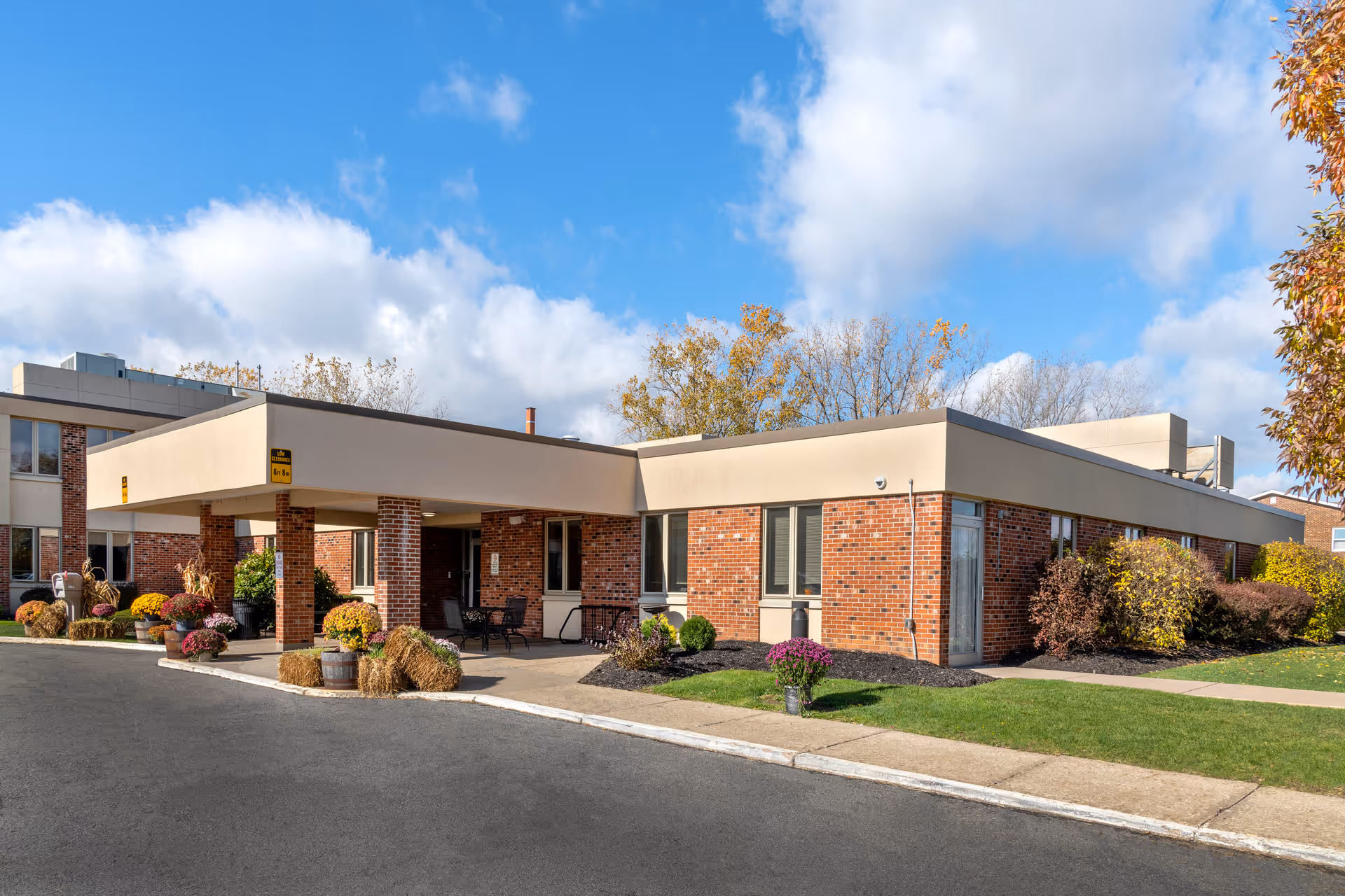 Exterior view of a single-story brick building with a covered entrance and a driveway. The building is surrounded by neatly maintained landscaping including bushes, flowers, and grass. The sky is blue with some clouds.
