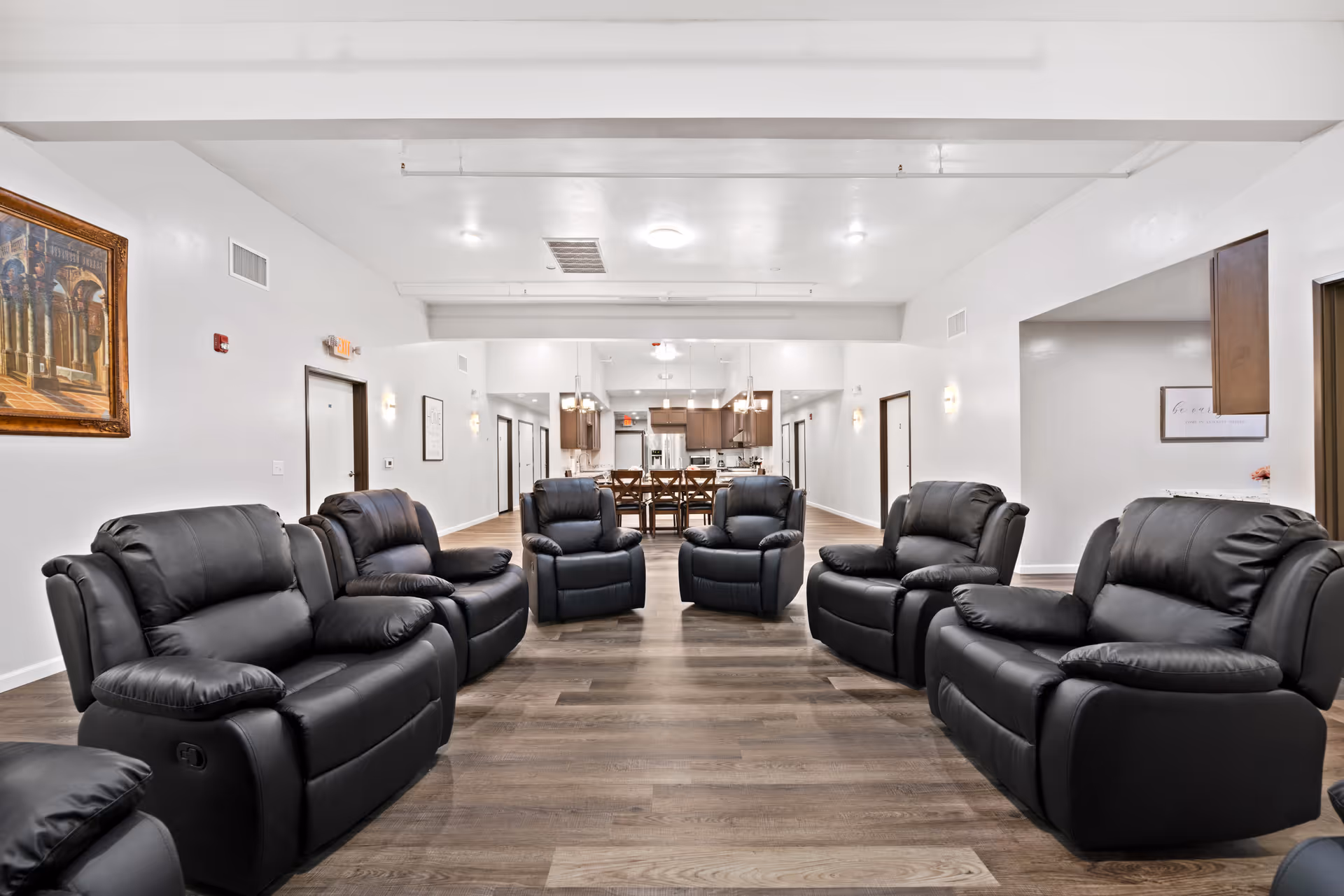 A spacious senior living common area with multiple black leather recliners arranged in a semi-circle on a wooden floor. The room has white walls, recessed lighting, and a large open kitchen and dining area visible in the background with wooden cabinets and a dining table with chairs.