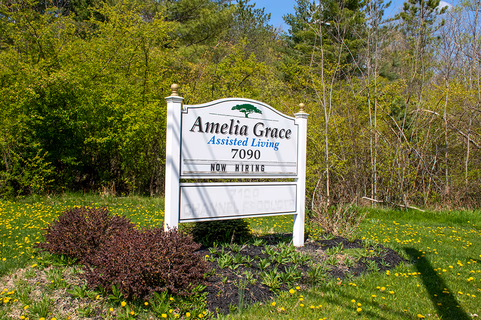 Outdoor view of a white sign for Amelia Grace Assisted Living with the address 7090, surrounded by green grass, yellow dandelions, bushes, and trees in the background under a clear blue sky.