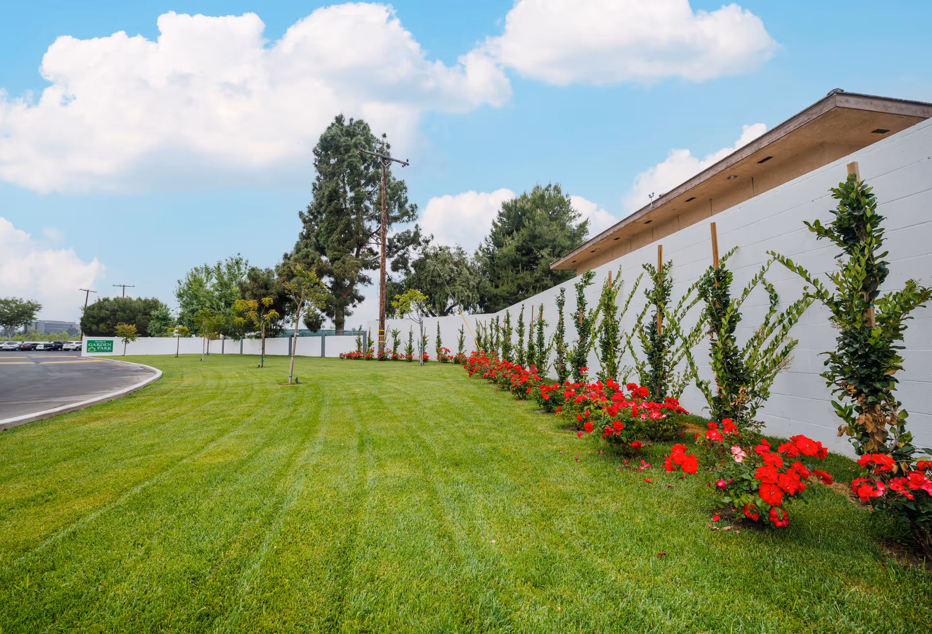 A well-maintained outdoor area with a green lawn, a row of small trees and red flowering plants along a white wall, under a partly cloudy blue sky. A curved driveway is visible on the left side with a green sign in the distance.