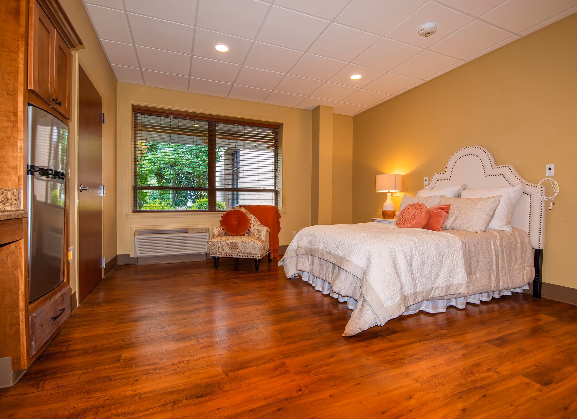 A cozy bedroom in Liberty Park Assisted Living featuring a large bed with multiple pillows and a white upholstered headboard. There is a bedside table with a lamp emitting warm light. A patterned armchair with an orange throw blanket and round pillow is placed near a window with wooden blinds, showing greenery outside. The room has wooden flooring and beige walls.