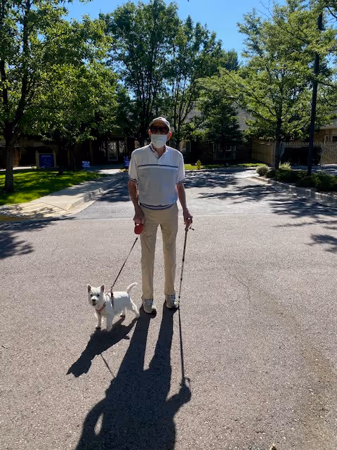 A person wearing a face mask walks a small white dog on a sunlit driveway lined with trees and buildings.