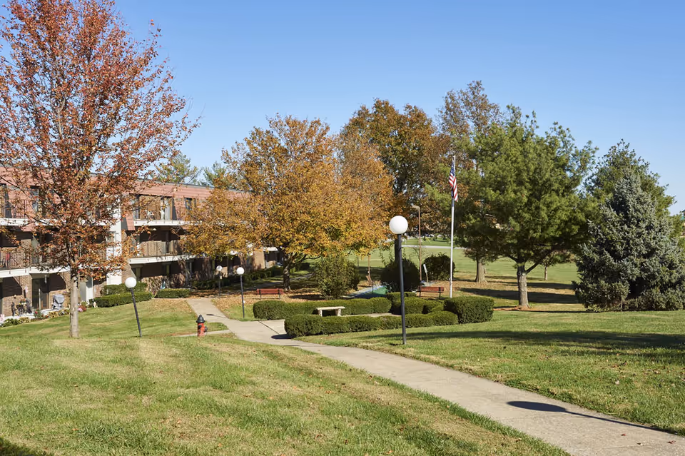 Outdoor view of a senior living facility with a paved walkway, green grass, trimmed bushes, several trees with autumn foliage, benches, lamp posts, and an American flag. A three-story building with balconies is visible on the left side under a clear blue sky.