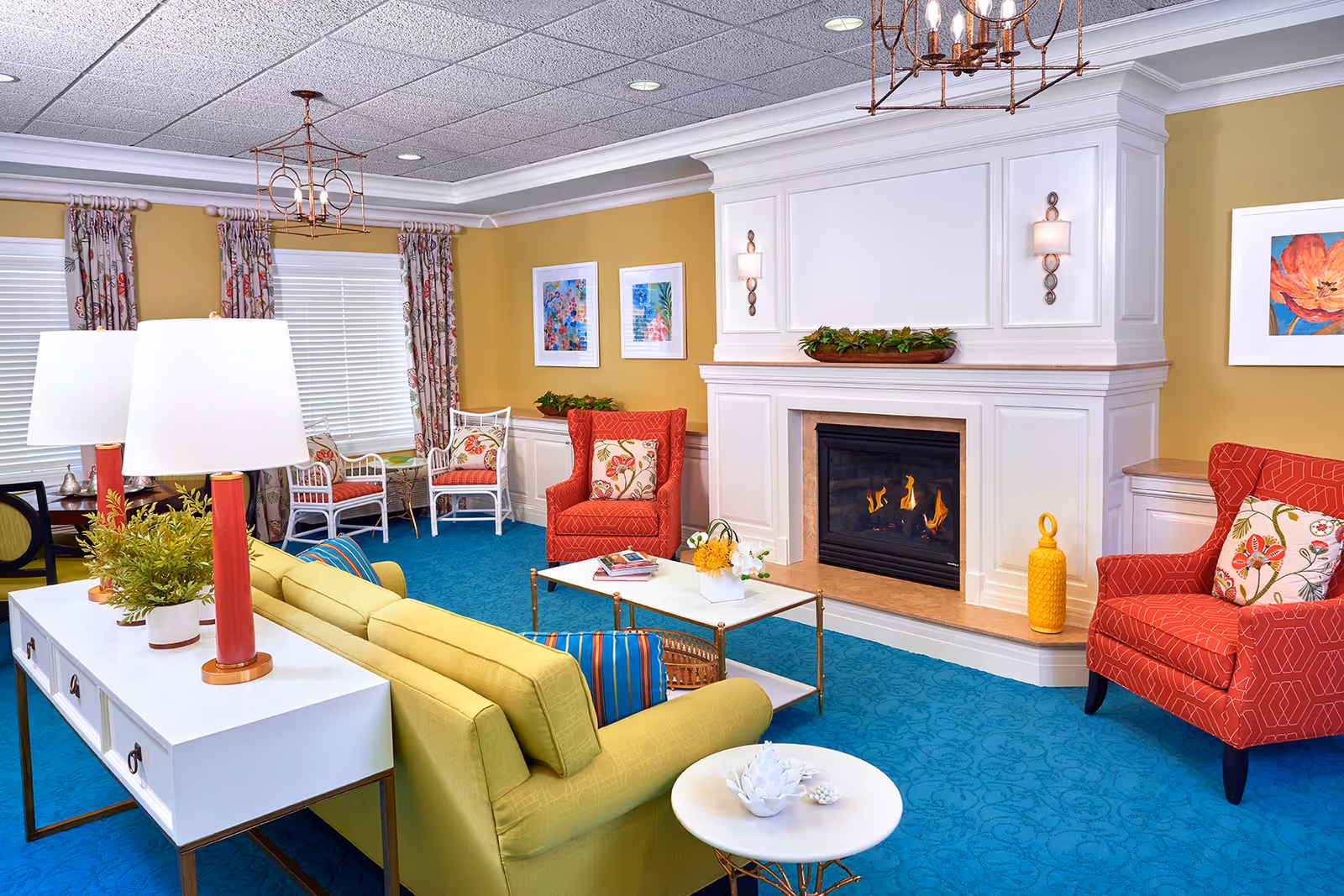 A cozy living room with a yellow sofa, two red armchairs with floral cushions, a white coffee table with flowers and books, a fireplace with a white mantel, blue carpet, yellow walls, and floral curtains on the windows.