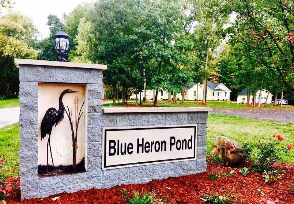 Stone sign for Blue Heron Pond with a metal sculpture of a blue heron and cattails on the left side, surrounded by mulch, flowers, and greenery with trees and houses in the background.