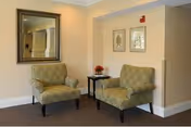 Two patterned upholstered chairs flank a small side table with a flower arrangement in a neutral-toned common sitting area with a mirror and framed art on the wall.