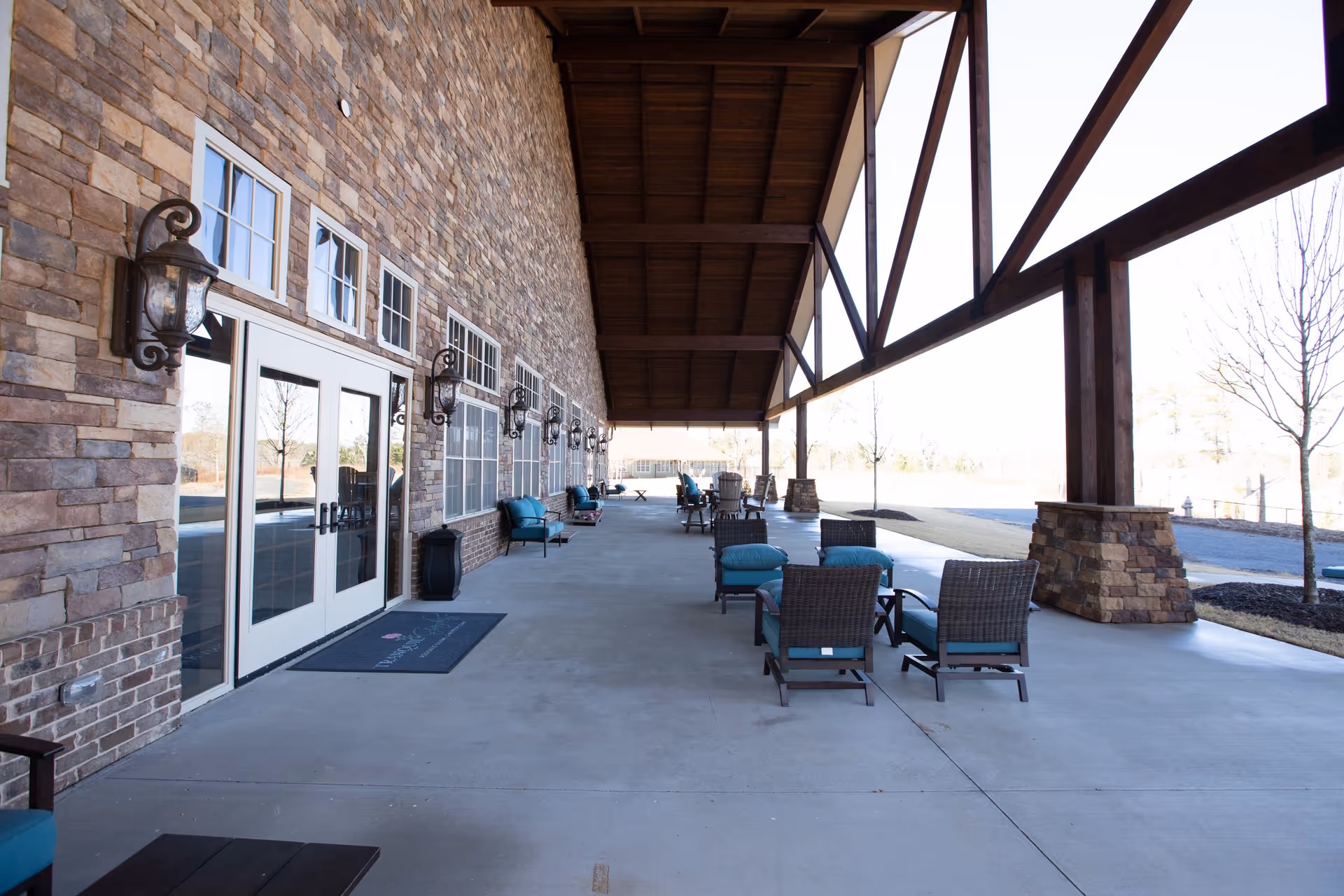 Covered outdoor patio with wicker chairs and teal cushions along a stone building facade and large wooden beams.