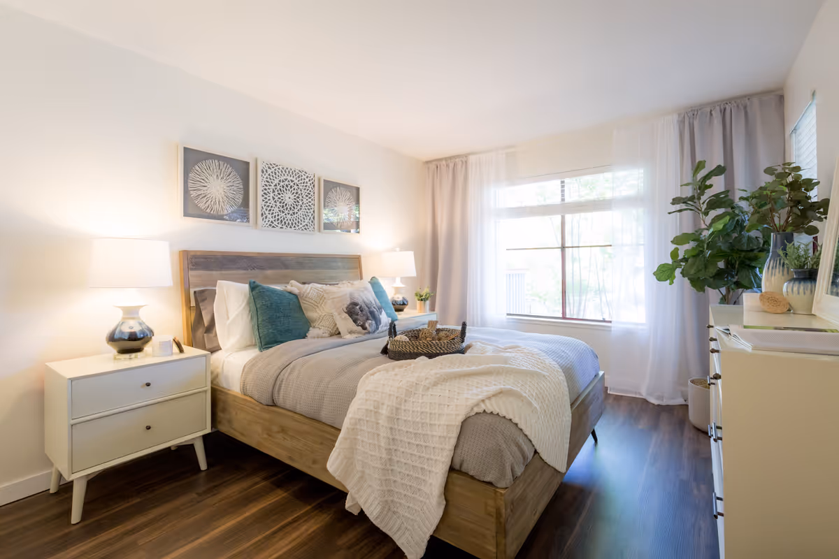Bright, modern bedroom with a wooden bed, bedside tables and lamps, decorative pillows and wall art, and a large window with sheer curtains.