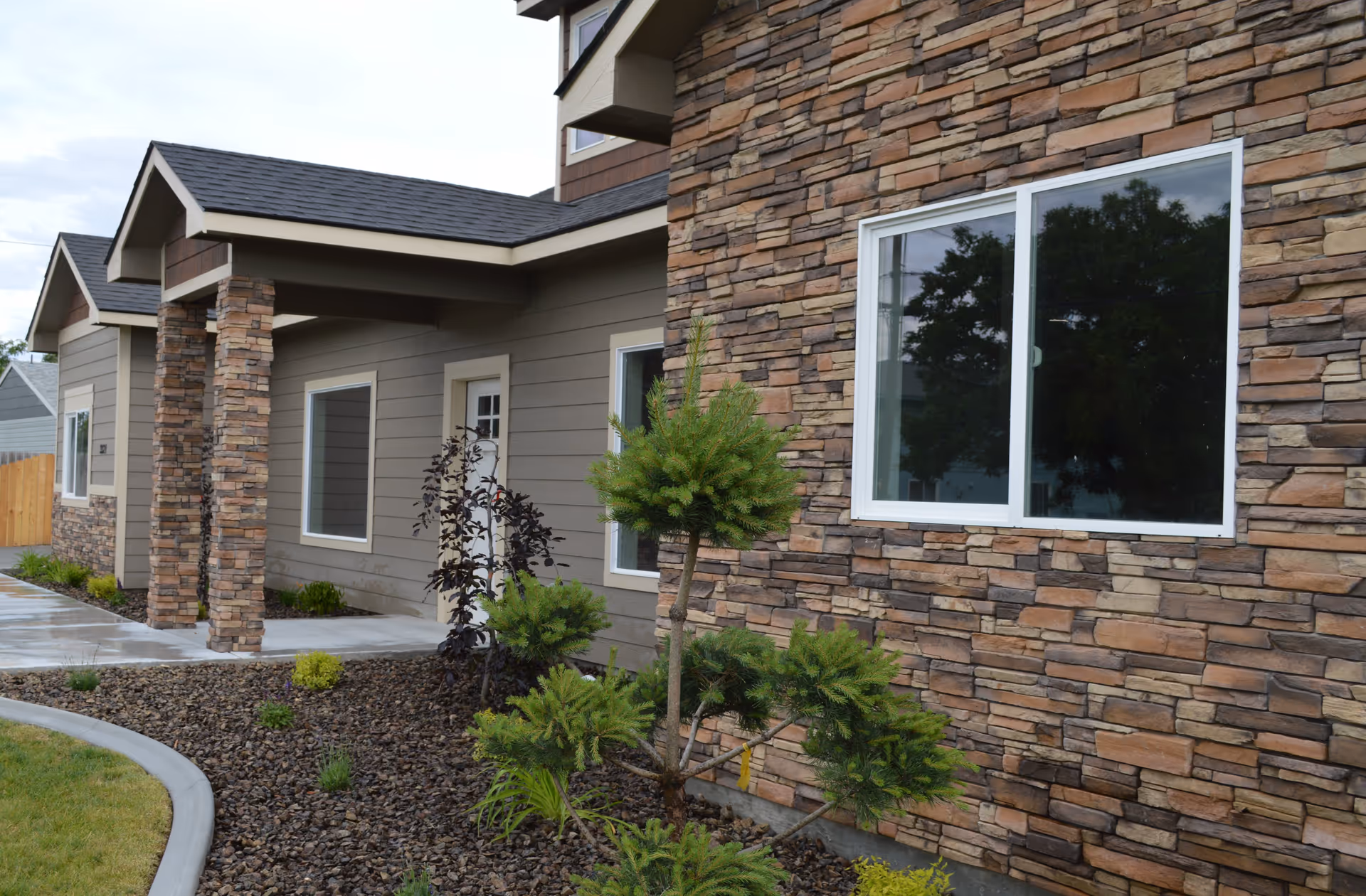 Exterior view of a building with stone and beige siding, featuring a covered entrance supported by stone pillars, several windows, and landscaping with small trees and shrubs along a curved concrete walkway.