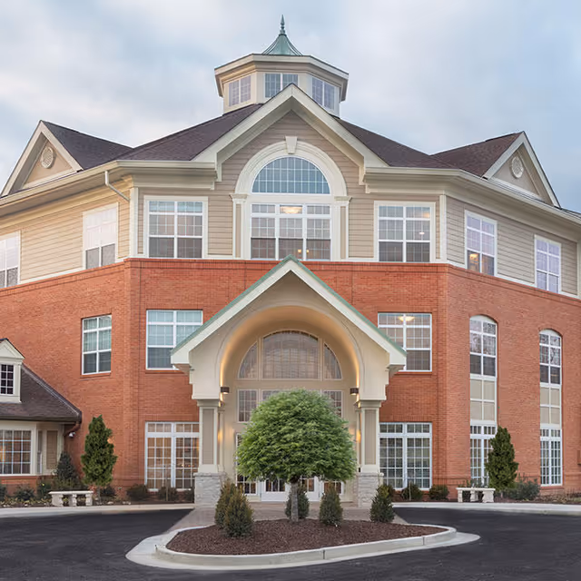 Front exterior view of a multi-story assisted living and memory care facility with a brick and beige facade, large windows, a peaked entrance with columns, and landscaped greenery including a small tree and shrubs in front.