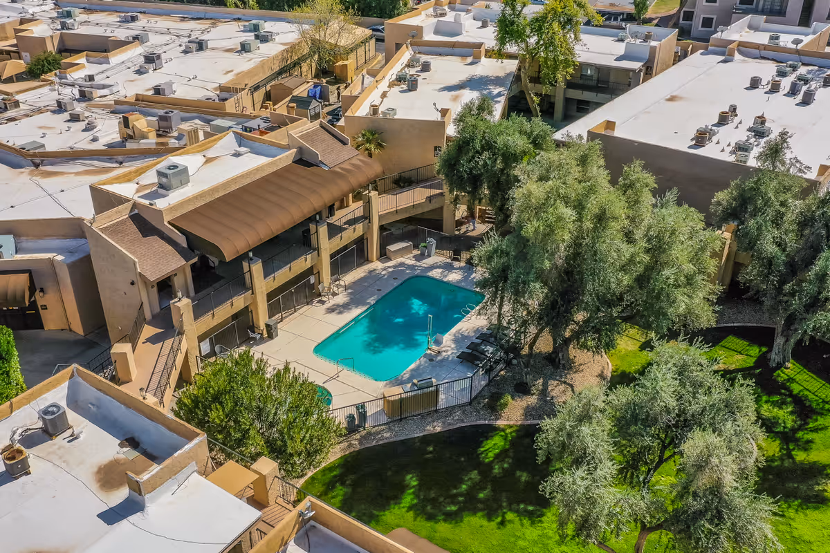 Aerial view of Scottsdale Village Square showing a swimming pool surrounded by a fenced patio area with lounge chairs, adjacent to a two-story building with balconies. The area is landscaped with green grass and several trees.