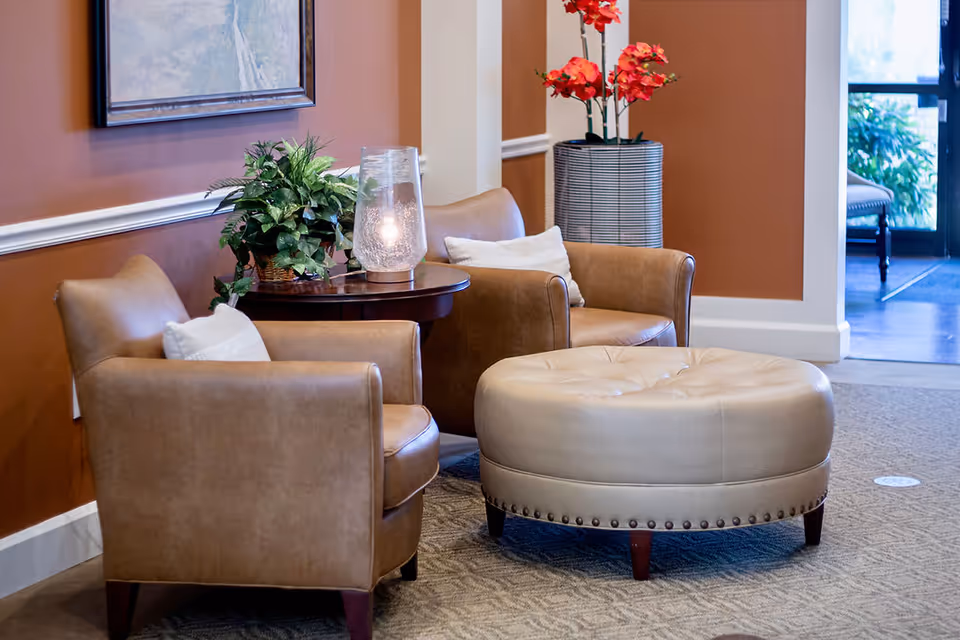 A cozy seating area with two tan leather armchairs, a round beige ottoman, a small wooden side table with a glass lamp and a green plant, and a tall vase with red flowers against a warm brown wall with white trim.