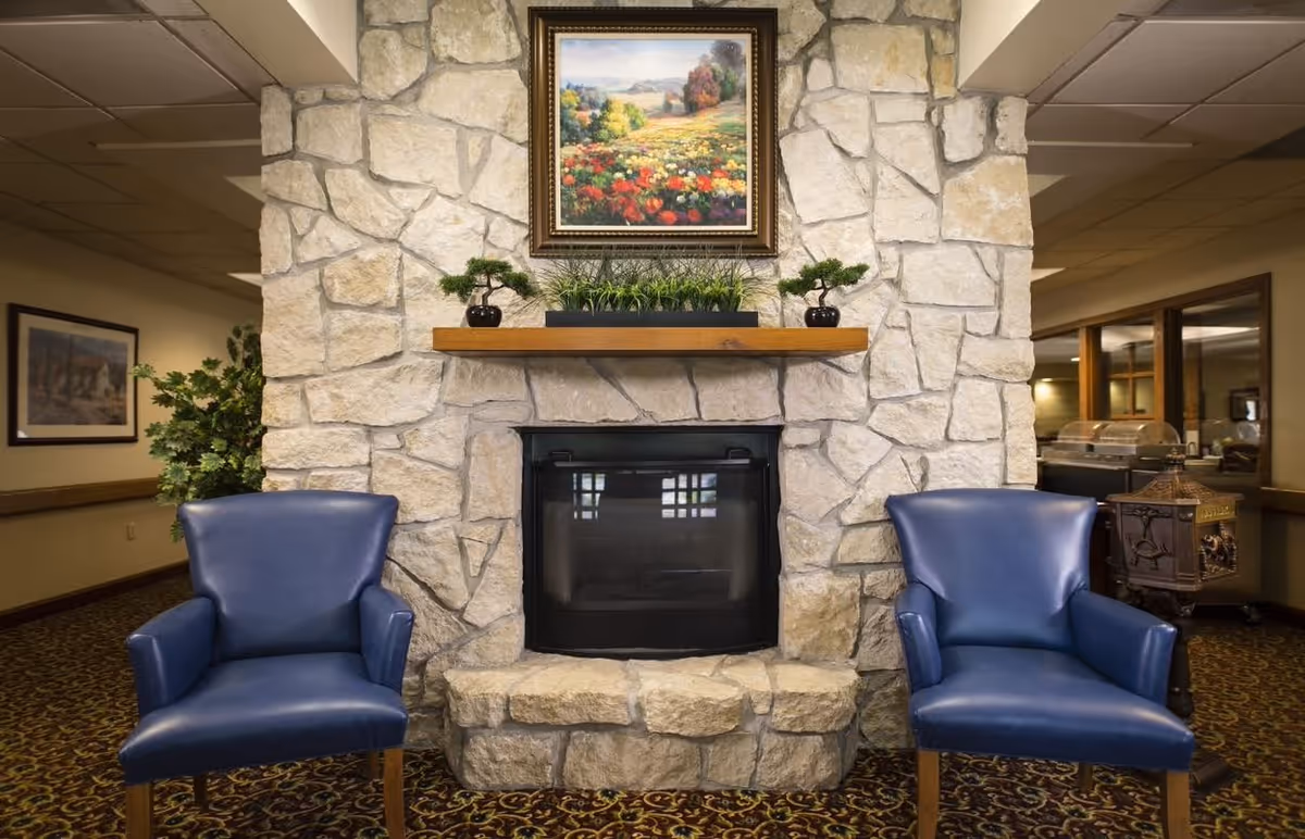 Stone fireplace with a wooden mantel and framed painting above, flanked by two blue armchairs in a communal lounge.