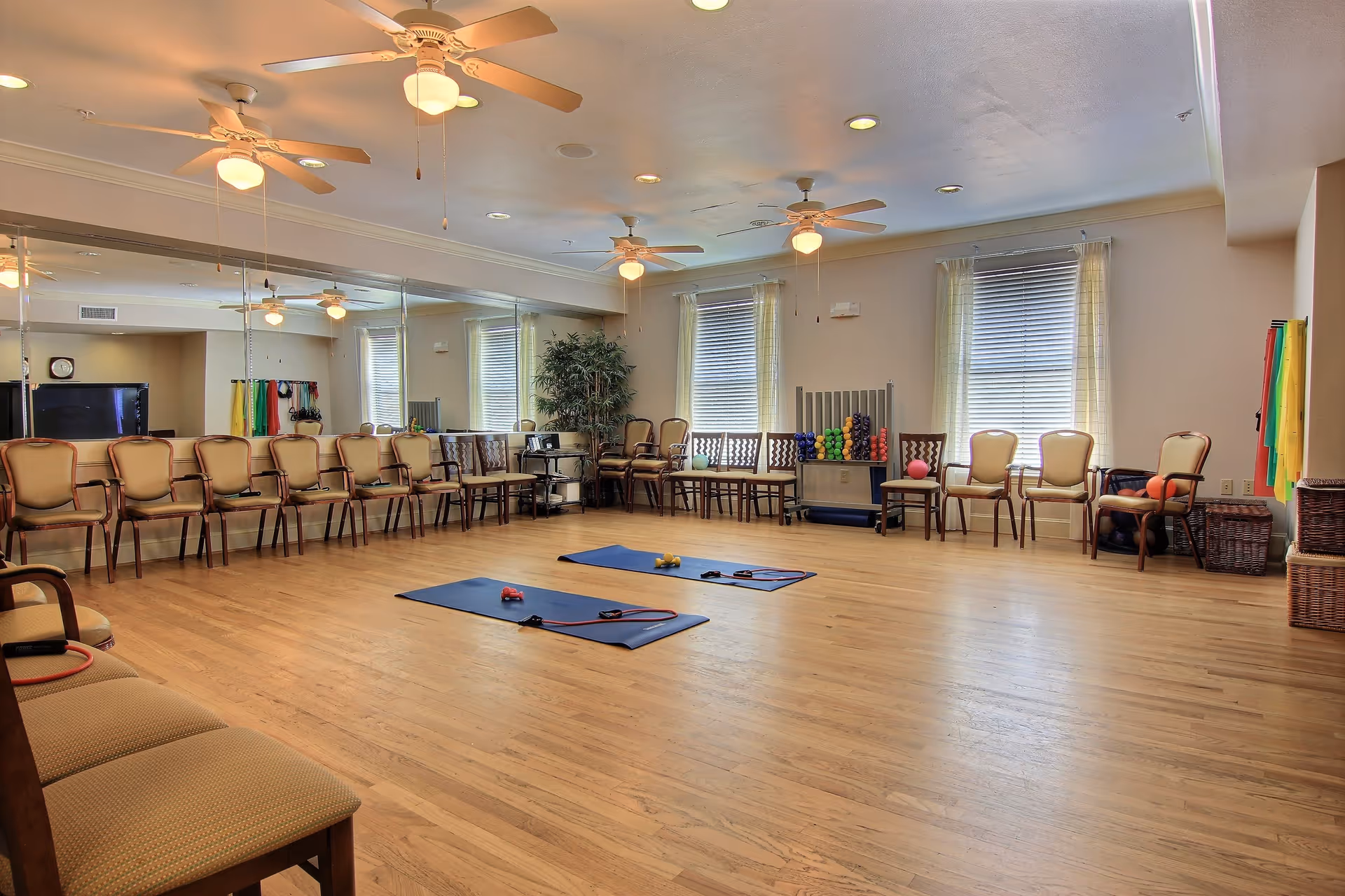 A spacious exercise room with wooden flooring, two blue exercise mats with resistance bands and small weights on them, a row of chairs lined up against the walls, large mirrors on one side, ceiling fans, windows with blinds and sheer curtains, and shelves with colorful exercise balls and equipment.