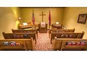 Interior view of a small chapel with wooden pews arranged in rows facing an altar with a cross on the wall behind it. The room is softly lit with lamps, and there are American and Christian flags on either side of the altar.