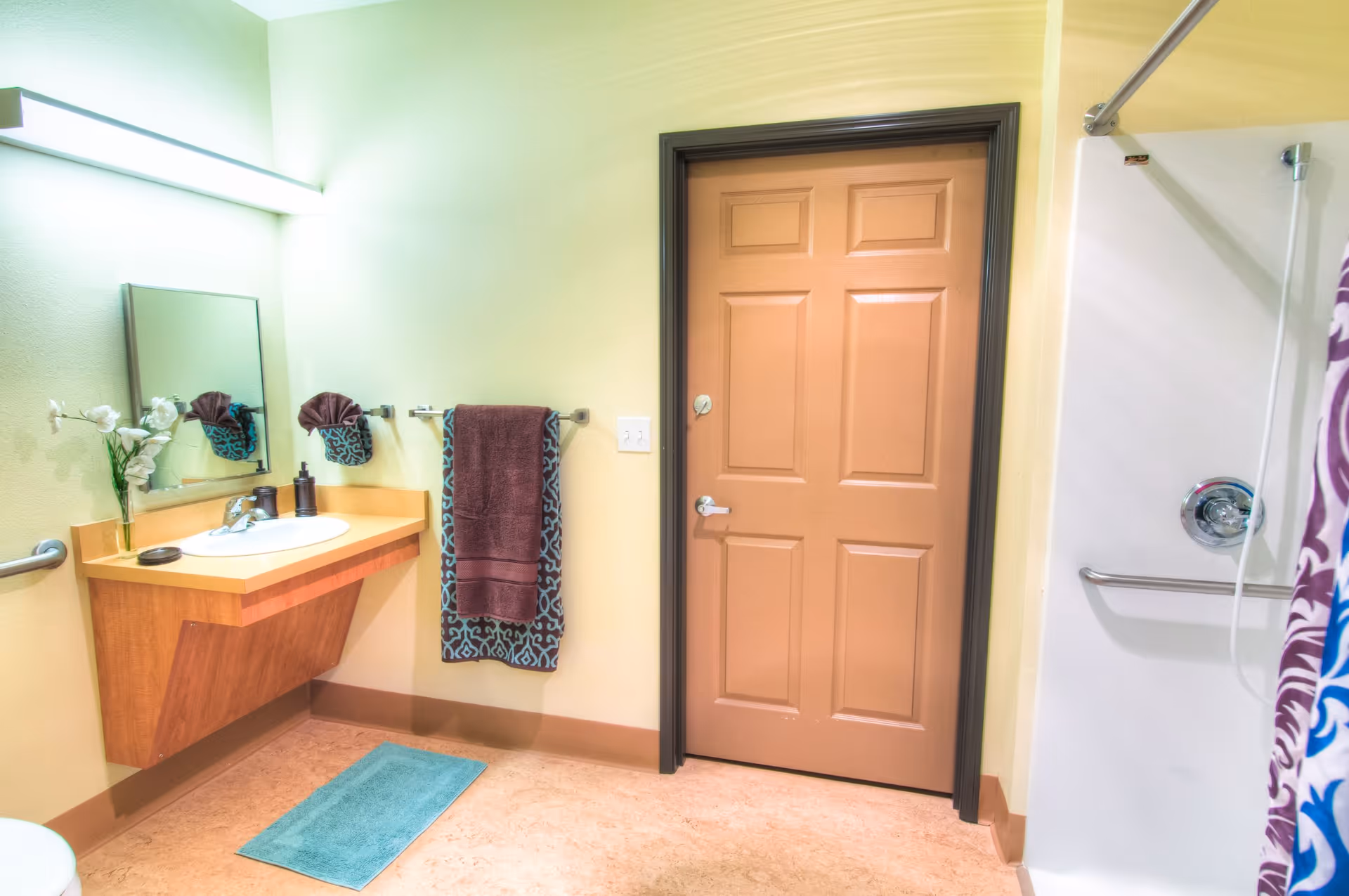 A clean and well-lit bathroom with a wall-mounted wooden sink vanity, a rectangular mirror above the sink, and a vase with white flowers on the counter. There are two towels hanging on the wall, one brown and one patterned. A closed brown door with a dark frame is visible, and to the right is a shower area with a grab bar and a colorful shower curtain.